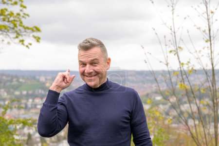 Photo for Middle age grey-haired attractive man wearing a blue sweater stands on a mountain and pointing to behind in the background you can see Stuttgart in Germany - Royalty Free Image