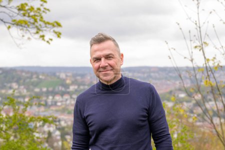 Photo for Middle age grey-haired attractive man wearing a blue sweater stands on a mountain and looks friendly to the camera in the background you can see Stuttgart in Germany - Royalty Free Image
