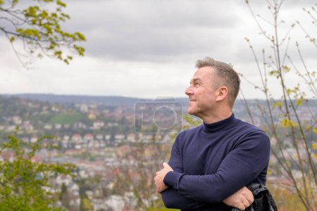 Foto de Hombre atractivo de pelo gris de mediana edad con un suéter azul se levanta en una montaña y mira pensativamente al lado en el fondo se puede ver Stuttgart en Alemania - Imagen libre de derechos
