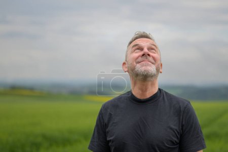 Photo for Middle age grey-haired man wearing a black shirt standing in front of a field and looking up what the weather ist doing - Royalty Free Image