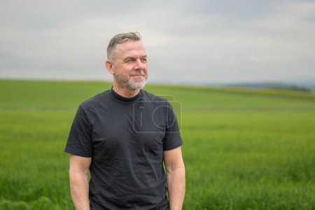 Téléchargez les photos : Homme aux cheveux gris du Moyen Âge portant une chemise noire debout devant un champ et semble amical sur le côté - en image libre de droit