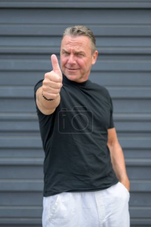Photo for Three quarter length portrait of a senior man showing thumbs up and looking to the camera against a blue wall - Royalty Free Image