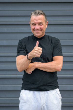 Photo for Three quarter length portrait of a senior man showing thumbs up and looking to the camera with focus to his face against a blue wall - Royalty Free Image