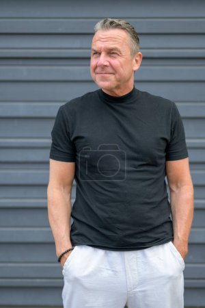 Photo for Three quarter length portrait of a senior man with both hands in his pants and looking to the side in front of a blue wall - Royalty Free Image