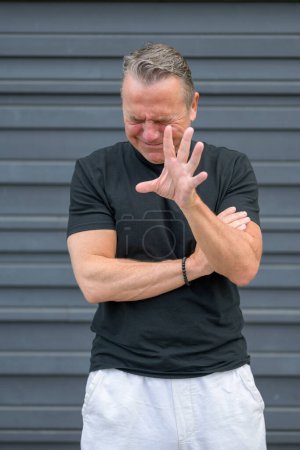 Téléchargez les photos : Portrait de trois quarts de long d'un homme âgé en détresse avec une main à la caméra disant "STOP" devant un mur bleu - en image libre de droit