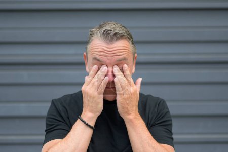 Foto de Primer plano de un hombre mayor frotándose los ojos porque estaba llorando o por alergia frente a una pared azul - Imagen libre de derechos
