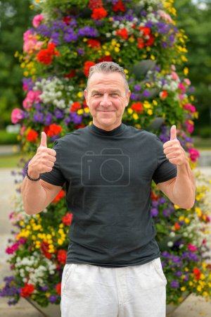 Photo for Three quarter length portrait of an attractive senior man showing both thumbs up in front of a large tower of flowers - Royalty Free Image