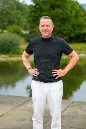 Foto de Retrato de tres cuartos de largo de un atractivo hombre mayor con las manos en las caderas frente a un pequeño lago - Imagen libre de derechos