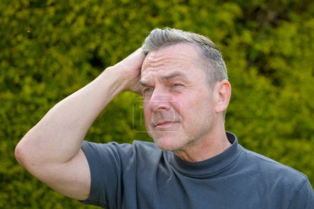 Foto de Retrato de un hombre de mediana edad con el pelo gris de pie en el jardín y mira a un lado y acariciando su cabello - Imagen libre de derechos