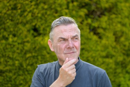 Foto de Retrato de un hombre de mediana edad con el pelo gris de pie en el jardín y mira hacia abajo con el dedo a la barbilla - Imagen libre de derechos