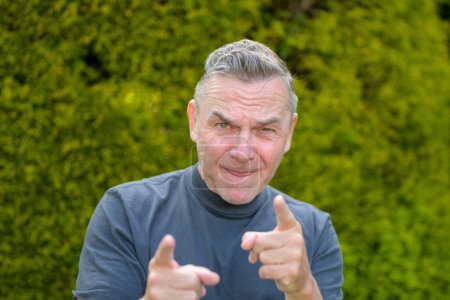 Foto de Retrato de un hombre de mediana edad con el pelo gris de pie en el jardín y señalando con dos dedos a la cámara - Imagen libre de derechos