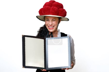 Photo for Close up portrait of a waitress in a traditional Bollenhut with red pompoms who winks mischievously and holding open a blank menu with copy space with a friendly smile - Royalty Free Image