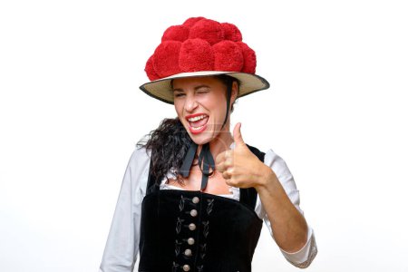 Téléchargez les photos : Enthousiaste riant jeune femme de la Forêt Noire portant une tradition bollenhut avec pompons rouges donnant un pouce geste d'approbation isolé sur blanc - en image libre de droit