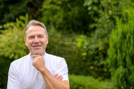 Foto de Atractivo hombre de mediana edad en sus 50 años sonriendo y con una mano en la barbilla mientras está de pie en el jardín - Imagen libre de derechos