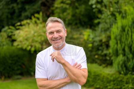 Foto de Atractivo hombre de mediana edad en sus 50 años sonriendo felizmente a la cámara mientras está de pie en el jardín - Imagen libre de derechos