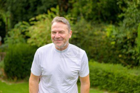Foto de Atractivo hombre de mediana edad en sus 50 años sonriendo a la cámara con una sonrisa traviesa mientras está de pie en el jardín - Imagen libre de derechos
