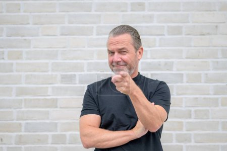 Téléchargez les photos : Portrait d'un séduisant homme sérieux pointant vers la caméra devant un mur de briques blanches - en image libre de droit