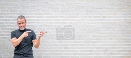 Photo for Promotional photo of an attractive serious man pointing to the right side in front of a white brick wall - Royalty Free Image
