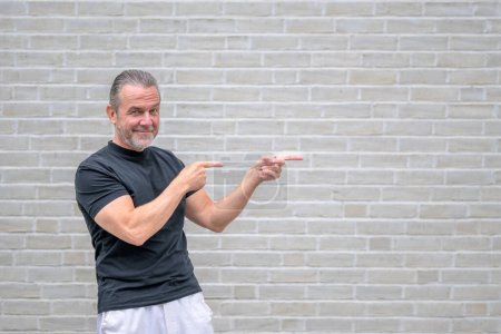 Photo for Portrait of an attractive friendly man pointing to the right side in front of a white brick wall - Royalty Free Image