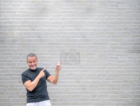Téléchargez les photos : Photo promotionnelle d'un homme sympathique attirant pointant vers le côté droit et devant un mur de briques blanches - en image libre de droit