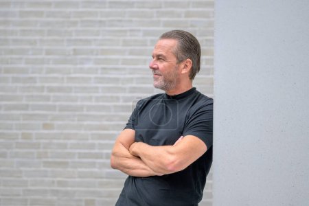 Foto de Hombre atractivo de pelo gris se apoya contra una pared con los brazos cruzados y mira pensativamente a la distancia delante de una pared de ladrillo blanco - Imagen libre de derechos