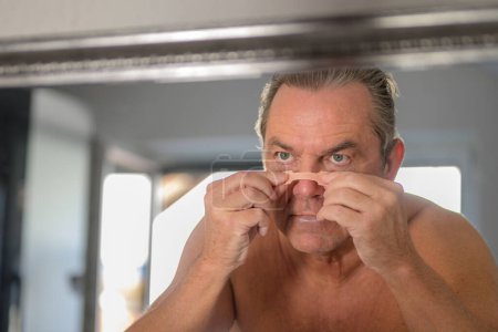 Foto de Hombre de mediana edad aplicando una cinta adhesiva mientras se mira en el espejo en el dormitorio - Imagen libre de derechos