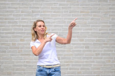 Photo for Close up portrait of an attractive friendly woman pointing to the right side and up in front of a white brick wall - Royalty Free Image