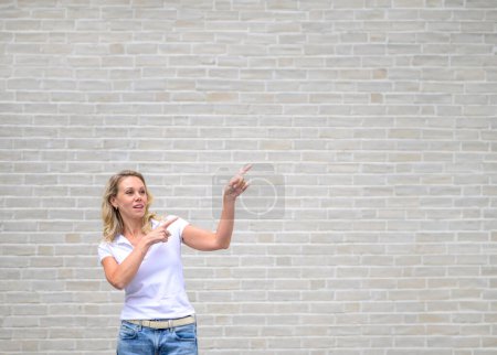 Photo for Promotional photo of an attractive friendly woman pointing to the right side and up in front of a white brick wall - Royalty Free Image