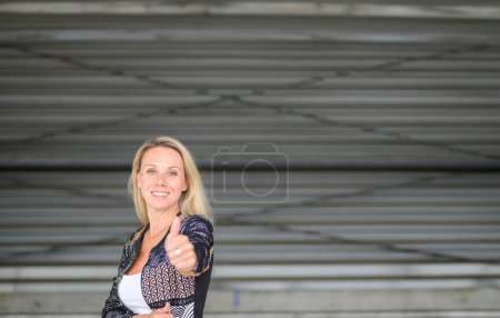 Foto de Retrato en la parte superior del cuerpo de una atractiva mujer rubia haciendo un gesto de pulgar hacia arriba con espacio para copiar, frente a una pared de hierro corrugado - Imagen libre de derechos