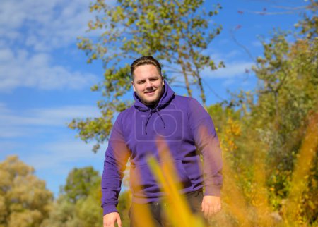 Foto de Retrato de la parte superior del cuerpo de un joven atractivo con una sudadera con capucha púrpura detrás de unos arbustos mirando a la cámara, afuera en otoño - Imagen libre de derechos