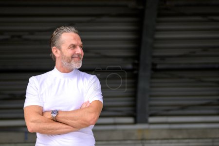 Photo for Side view of an attractive gray haired man with a white T-shirt has his arms crossed, wears a stylish watch, and looks friendly to the side, in front of a corrugated iron wall - Royalty Free Image