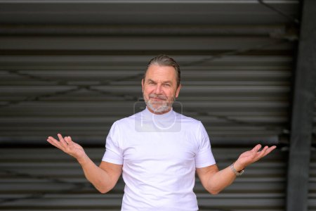 Foto de Atractivo hombre de pelo gris con una camiseta blanca tiene los brazos extendidos y mira cuestionablemente a la cámara, frente a una pared de hierro corrugado - Imagen libre de derechos