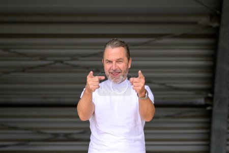 Téléchargez les photos : Charmant homme aux cheveux gris avec un T-shirt blanc portant une montre élégante et pointant malicieusement vers la caméra, devant un mur de fer ondulé - en image libre de droit