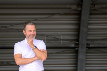 Photo for Side view of an attractive gray haired man with a white T-shirt has his arms crossed, wears a stylish watch, and looks critically to the side, in front of a corrugated iron wall - Royalty Free Image