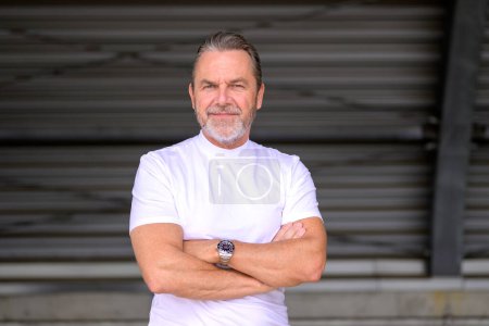 Foto de Atractivo hombre de pelo gris con una camiseta blanca tiene los brazos cruzados, lleva un reloj elegante, y se ve amigable a la cámara, frente a una pared de hierro corrugado - Imagen libre de derechos