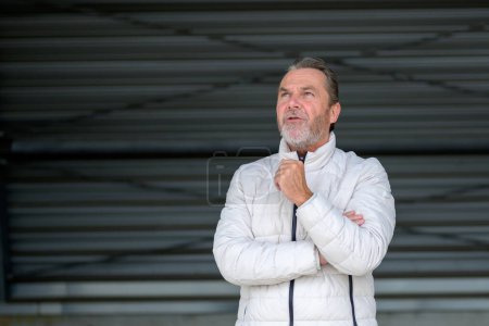 Téléchargez les photos : Charmant homme aux cheveux gris portant une veste d'hiver blanche avec une main sur sa veste regardant étonné vers le ciel, devant un mur de fer ondulé - en image libre de droit
