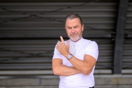 Téléchargez les photos : Homme aux cheveux gris attrayant avec un t-shirt blanc portant une montre de fantaisie et regardant de manière critique et demandant à la caméra, devant un mur de fer ondulé - en image libre de droit