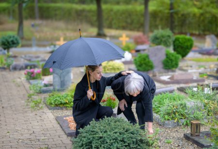 Téléchargez les photos : Fille étreignant sa mère à la tombe de l'homme dans le cimetière par une journée froide nuageuse tout en prenant soin de la tombe - en image libre de droit