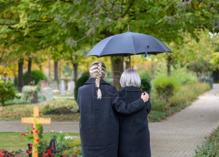 Téléchargez les photos : Vue arrière de la fille tenant sa mère dans ses bras sur le chemin de la tombe de l'homme dans le cimetière par une journée nuageuse et froide - en image libre de droit