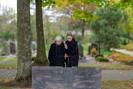 Téléchargez les photos : Mère et fille debout sur la tombe de l'homme dans le cimetière par une journée nuageuse froide et regardant vers le bas - en image libre de droit