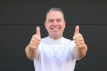 Téléchargez les photos : Un homme mûr souriant montrant ses nouvelles dents et donnant un double pouce devant un mur noir - en image libre de droit