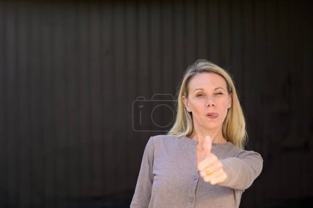 Photo for Portrait of an attractive winking woman sticking out her tongue giving a thumbs up gesture, in front of wooden wall - Royalty Free Image