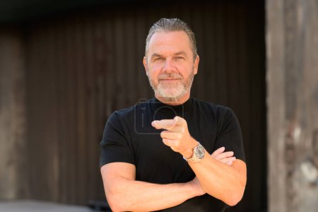 Téléchargez les photos : Attrayant homme aux cheveux gris avec un T-shirt noir porte une montre élégante et pointant vers l'appareil photo, en face d'un mur en bois - en image libre de droit
