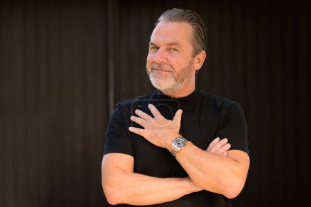 Charmant homme aux cheveux gris portant un T-shirt noir et montrant sa montre élégante, devant un mur en bois