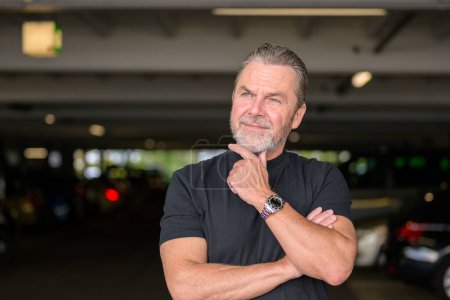 Foto de Atractivo hombre de pelo gris con una camiseta negra lleva un reloj elegante y mirando hacia un lado, delante de un aparcamiento - Imagen libre de derechos
