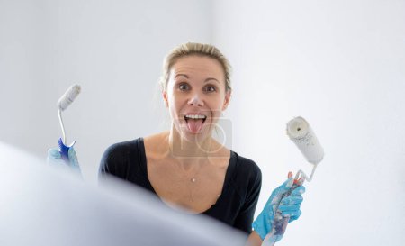 Photo for Close up of a beautiful blonde woman is having fun, sticking out her tongue while painting the apartment and holding two paint rollers in her hands and looking at the camera, in her new apartment - Royalty Free Image