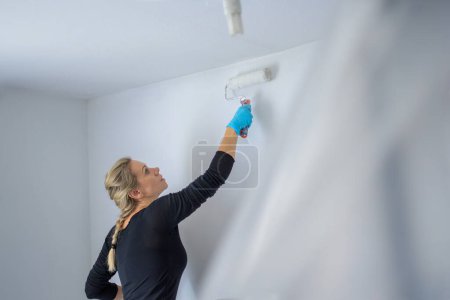 Téléchargez les photos : Femme célibataire blonde peignant le mur de son appartement avec un grand rouleau de peinture, dans son nouvel appartement - en image libre de droit