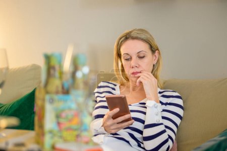 Photo for Attractive middle aged blonde woman sitting on the couch and using her smartphone - Royalty Free Image