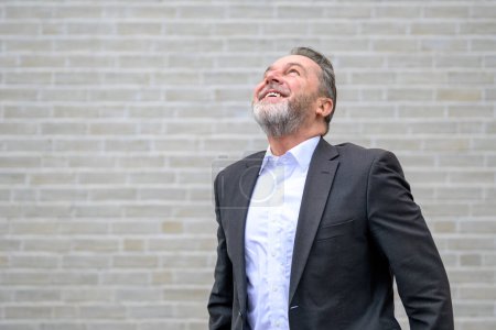 Foto de Atractivo hombre gris de sesenta años frente a una pared de ladrillo blanco mirando hacia arriba con una gran sonrisa - Imagen libre de derechos