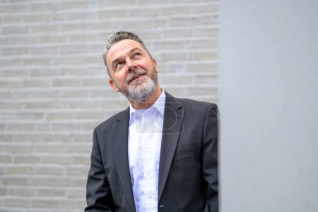 Foto de Atractivo hombre gris de sesenta años frente a una pared de ladrillo blanco mirando hacia arriba desde detrás de una cornisa con una sonrisa - Imagen libre de derechos